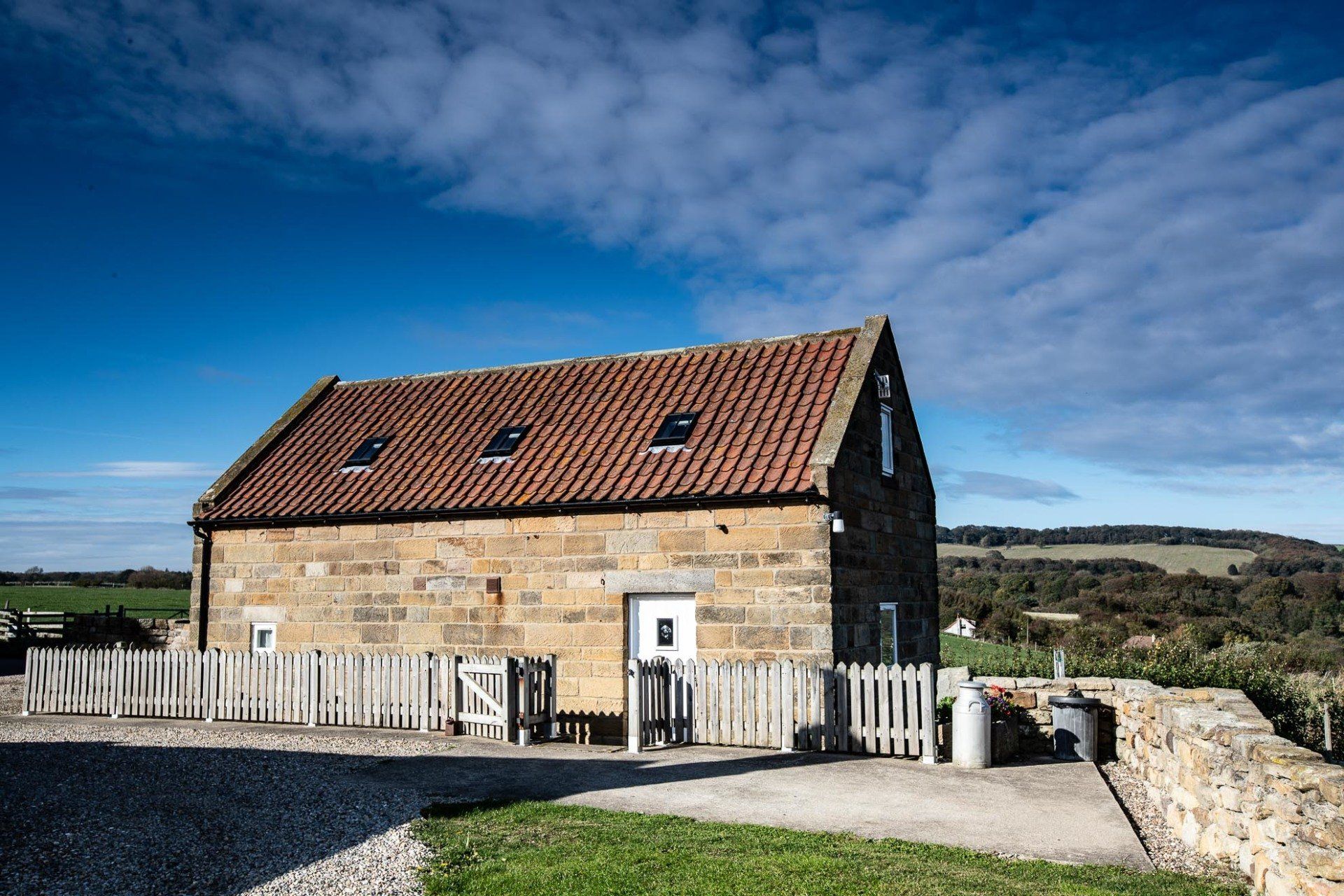 Stone cottage with red tile roof, white picket fence, and green lawn under a blue sky.