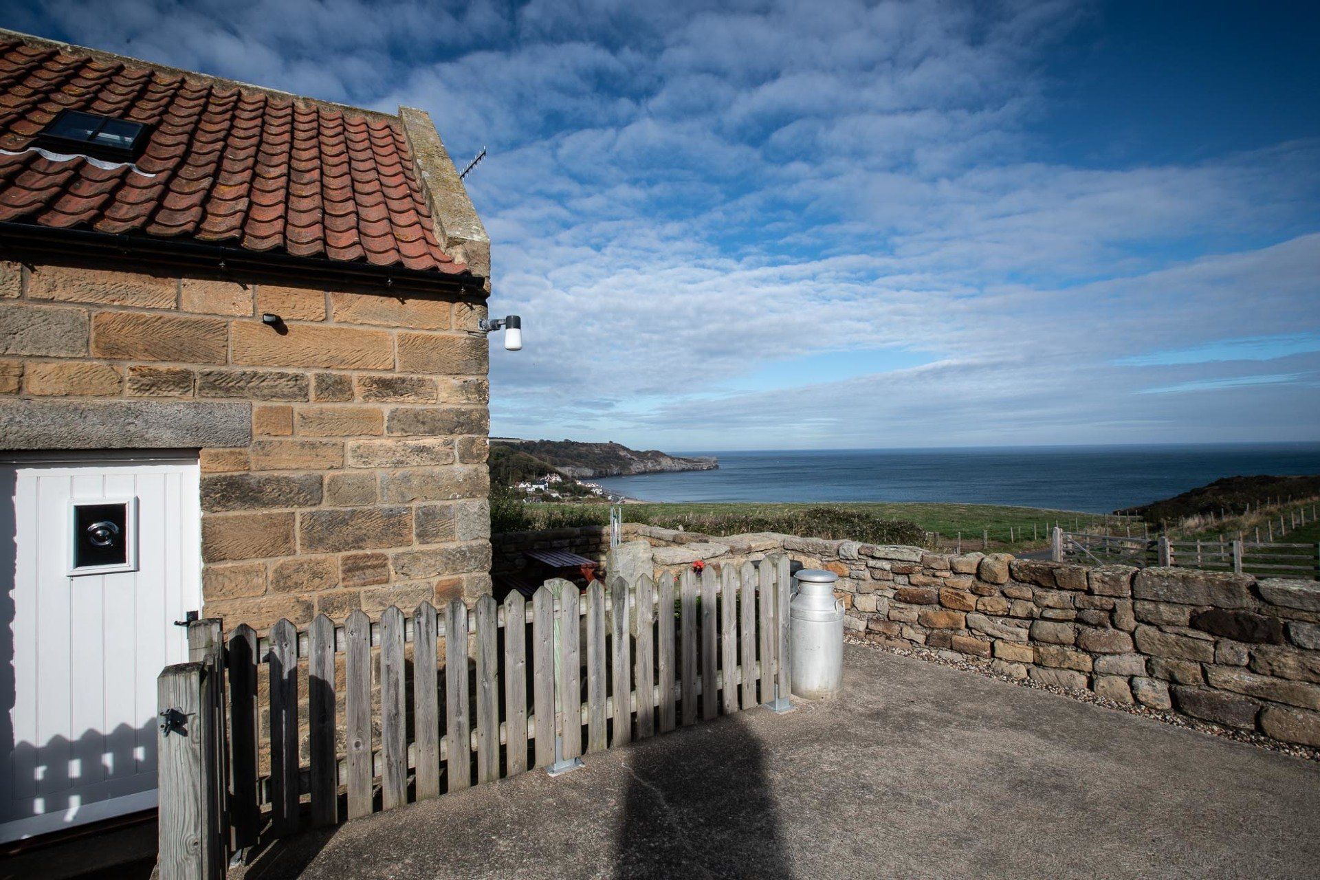 Stone cottage with a sea view. Brown tiled roof, white door, wooden fence, blue sky, and ocean.