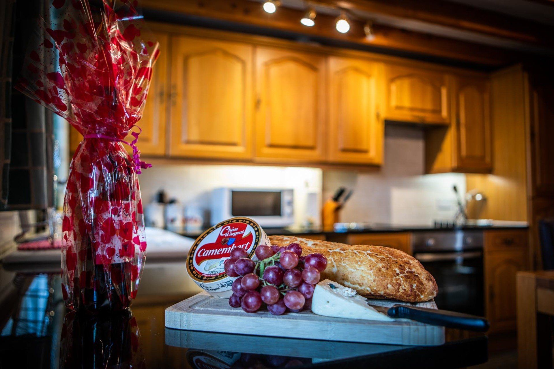 Kitchen still life: wine bottle, wrapped in red, with cheese, grapes, and bread on a cutting board.