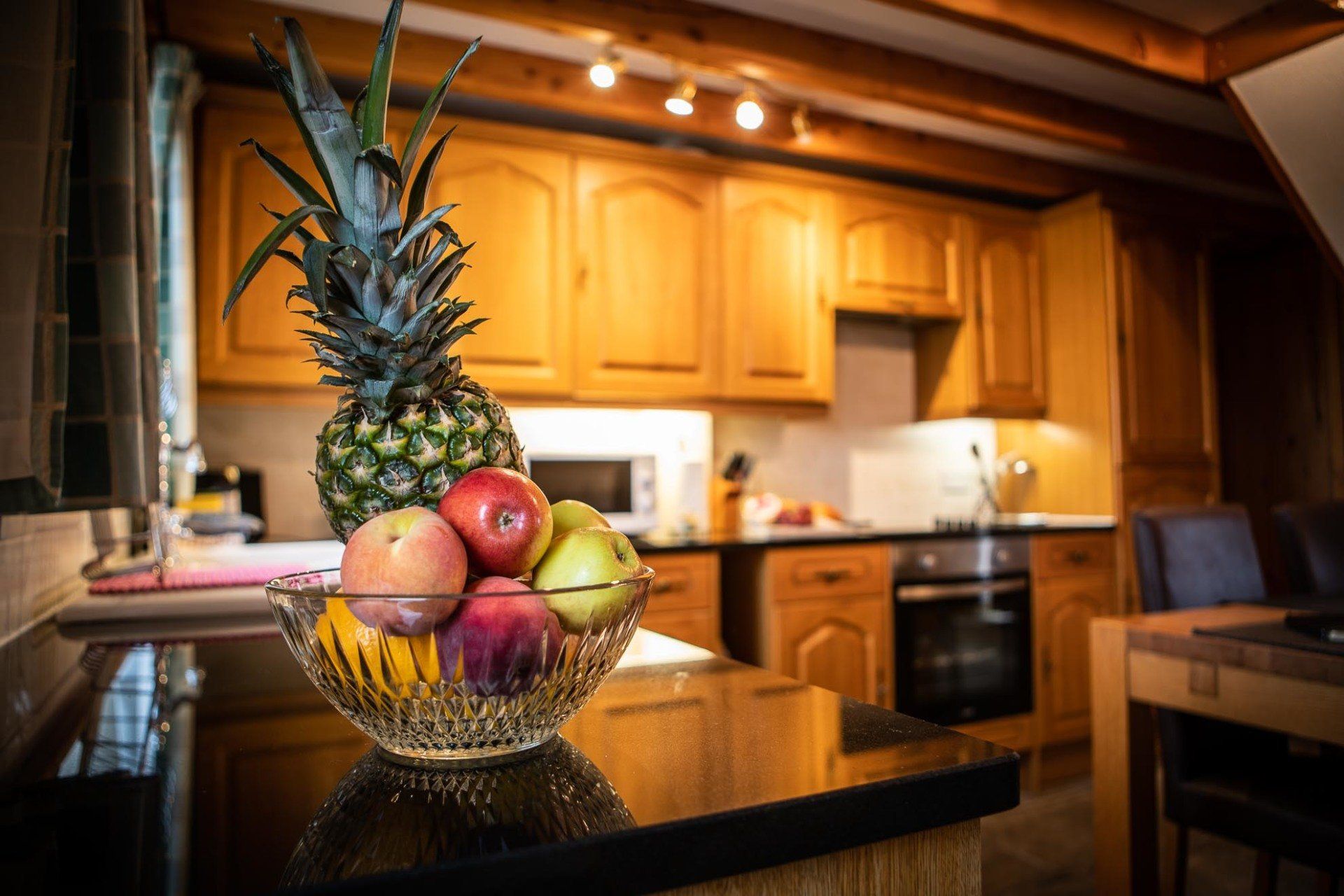 Fruit bowl with pineapple, apples, and peaches on a kitchen countertop; wooden cabinets and oven in background.