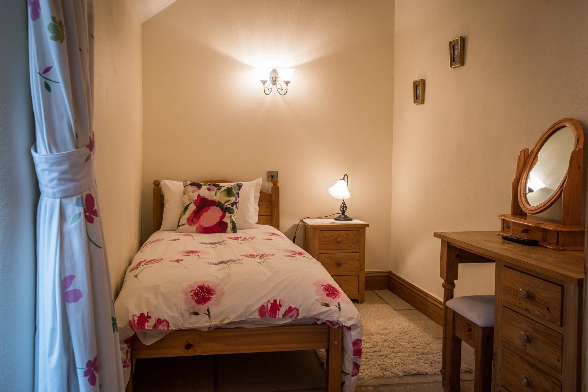 Small bedroom with single bed, dresser, and nightstand. Floral bedding and curtain, warm lighting.