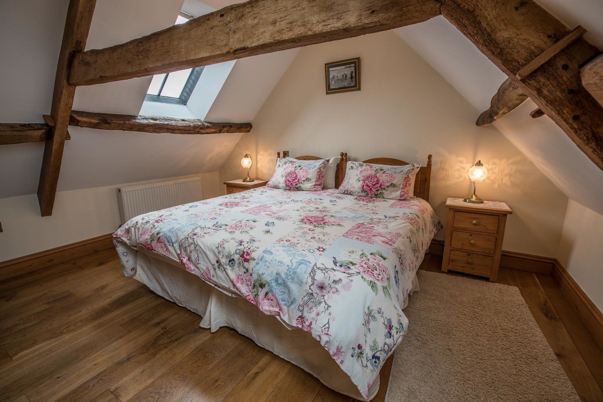 Bedroom in attic with bed, floral bedding, wooden beams, nightstands, and window.