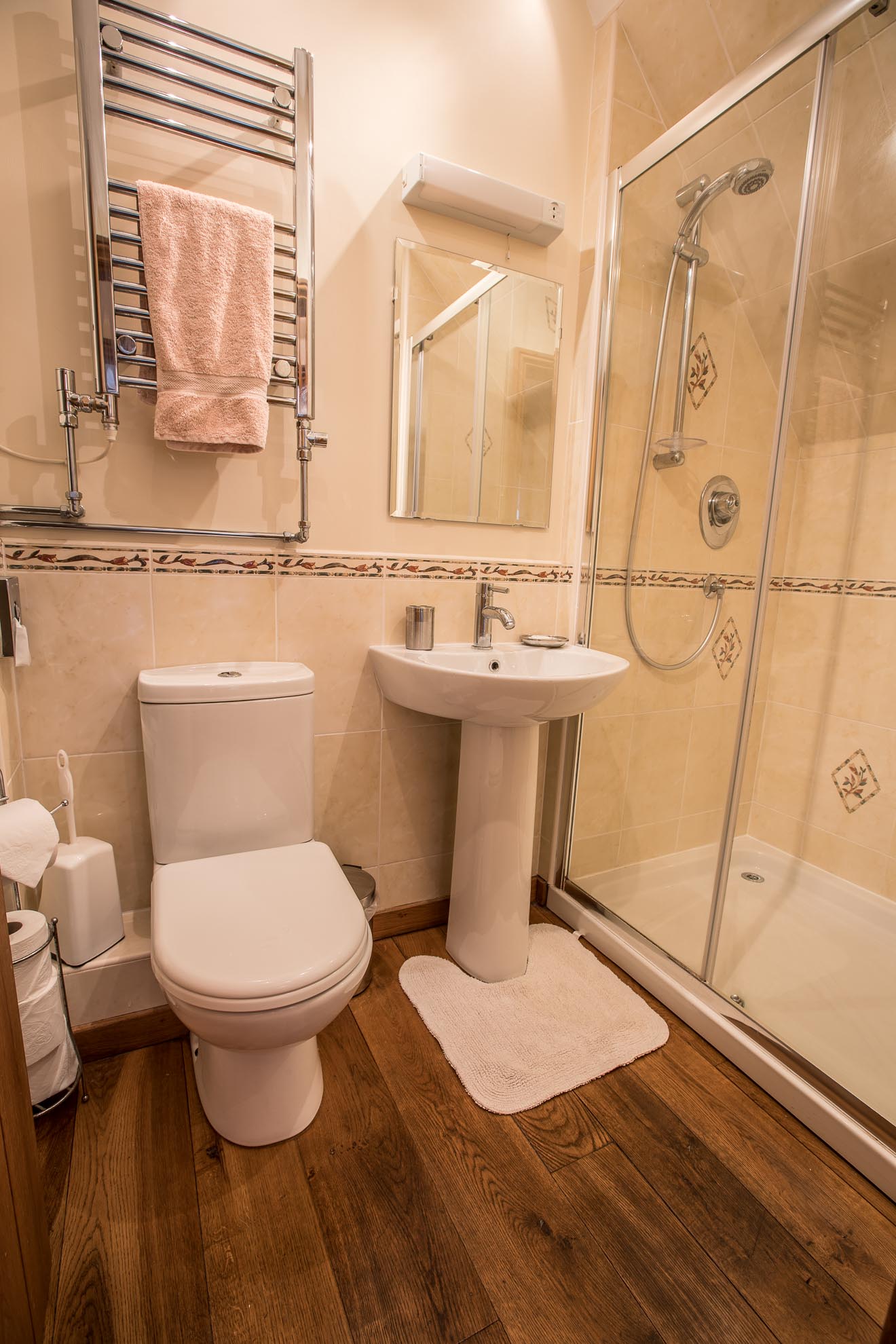 Bathroom with white toilet, sink, shower, and towel rack; wooden floor.