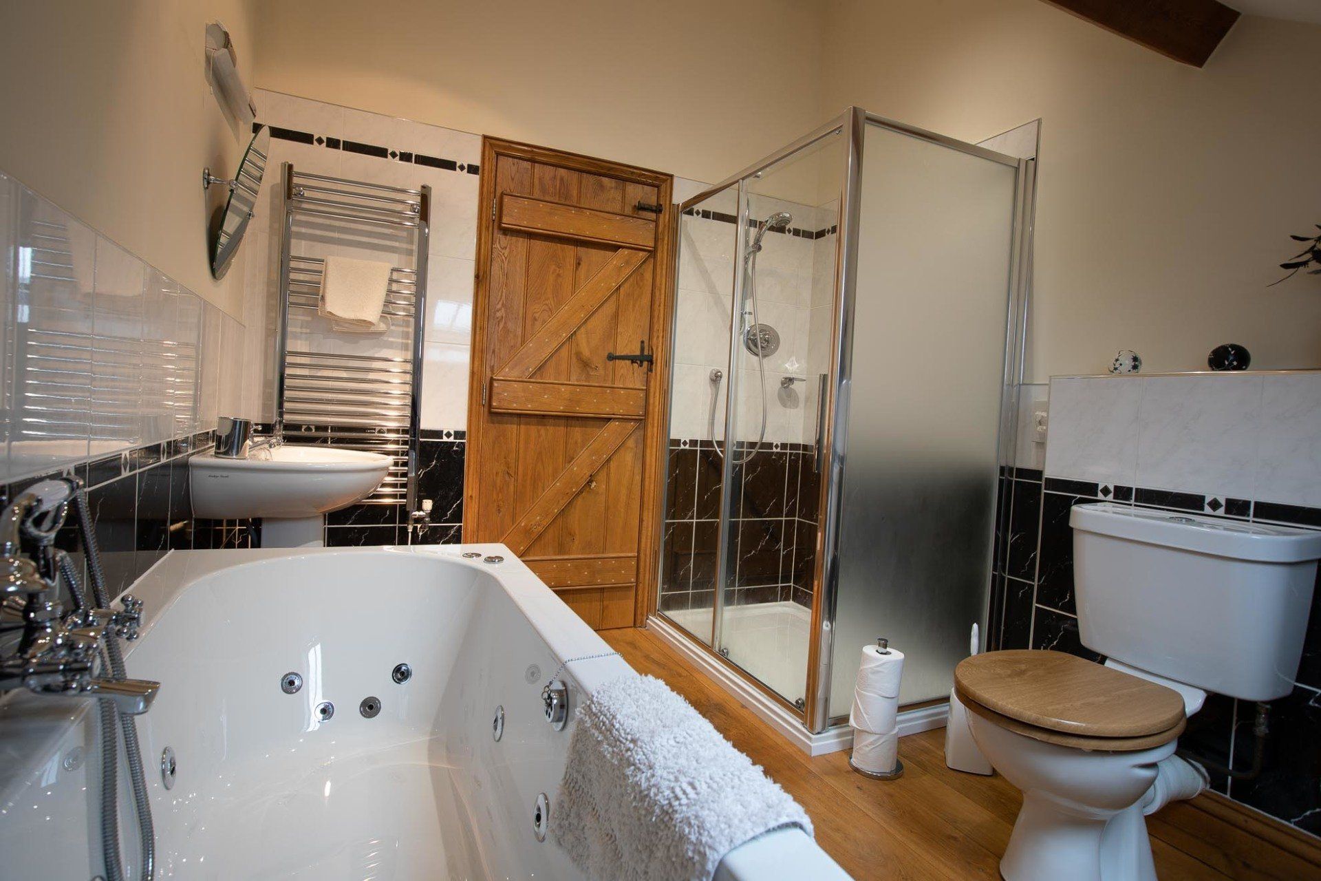 Bathroom with tub, shower, and toilet; black and white tiles, wooden door, and a towel on the tub's edge.