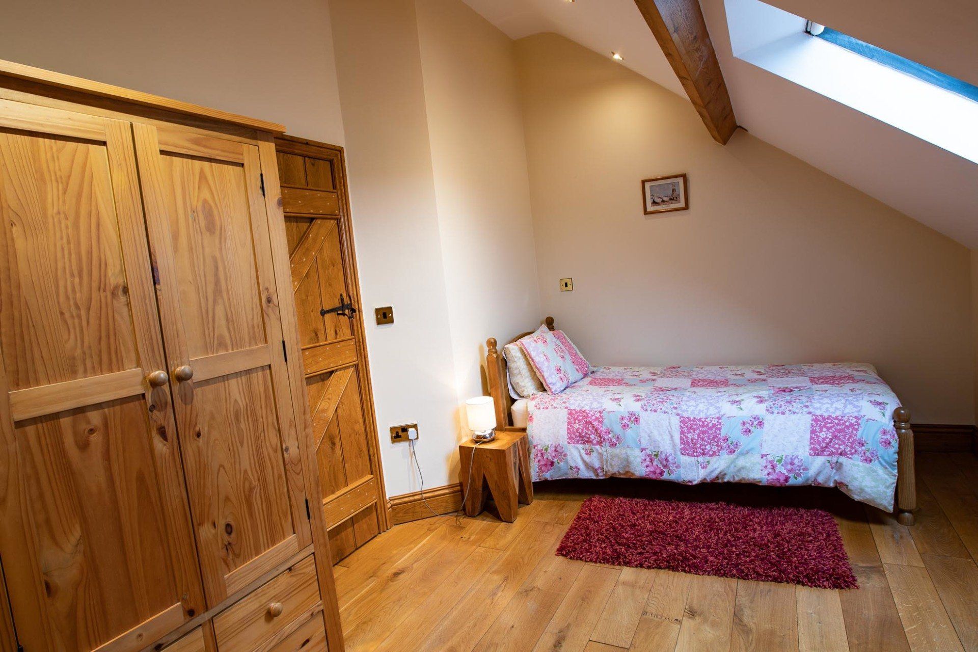 Bedroom with wooden furniture, including a wardrobe, bed with pink bedding, and small rug. Natural light from a skylight.
