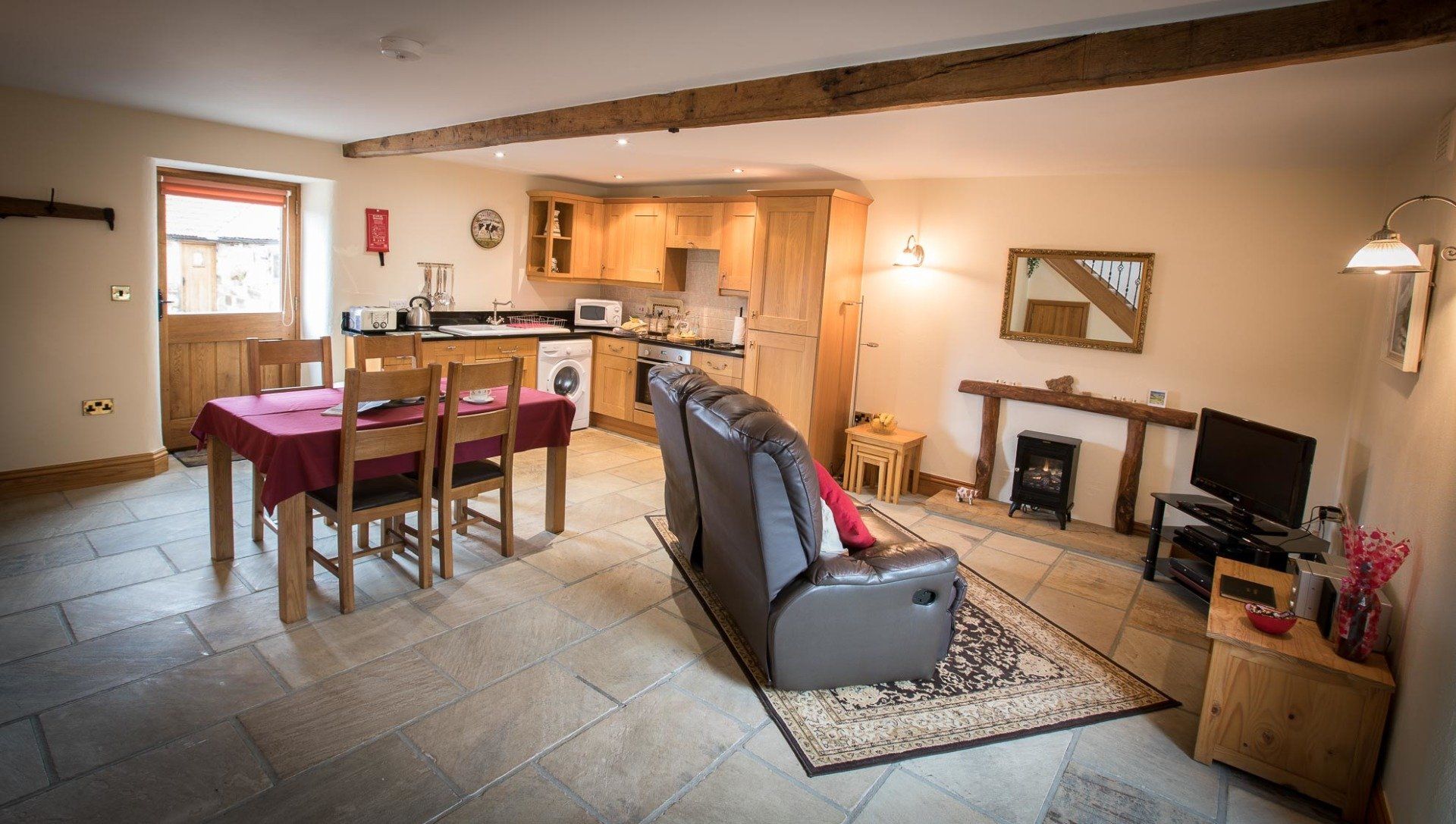 Cozy cottage interior: kitchen, dining area, living room with fireplace, brown beams, gray stone floor, and a leather recliner.