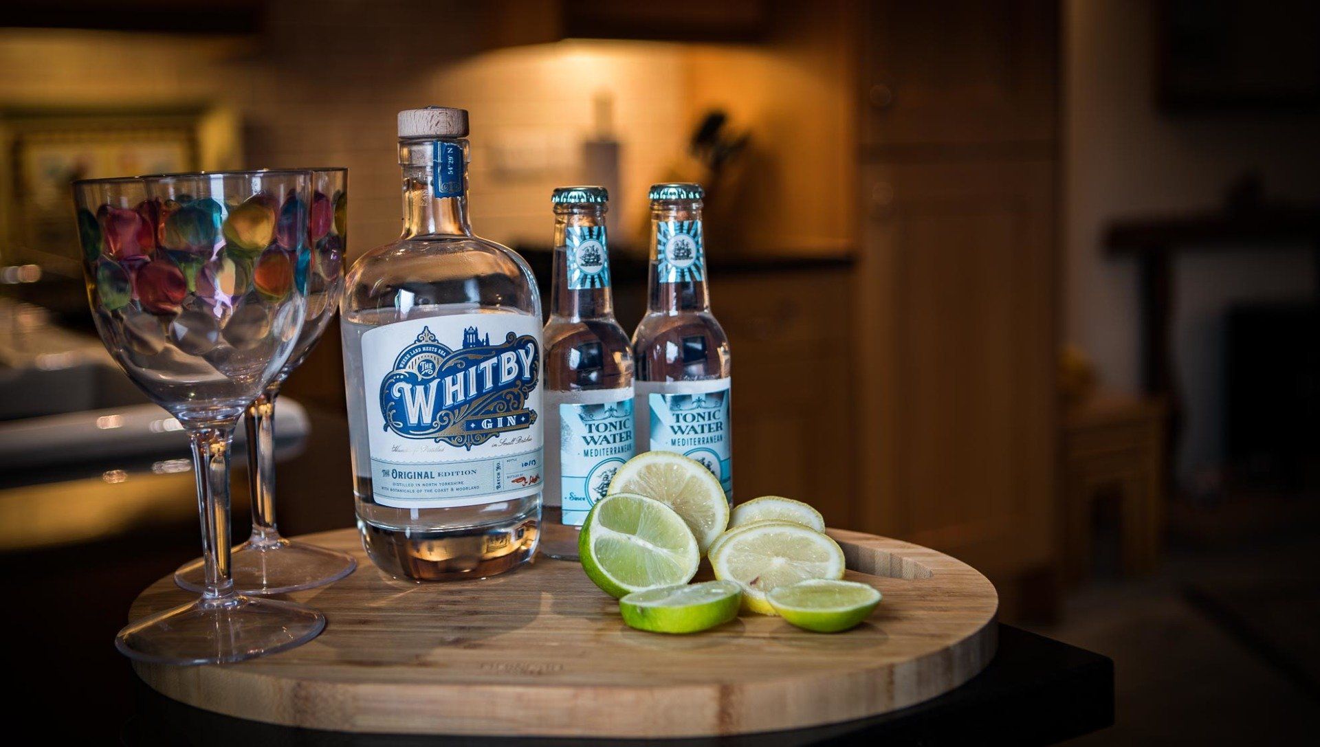 Gin and tonics ingredients on wooden tray. Bottle, two mixers, sliced limes, glass. Kitchen background.