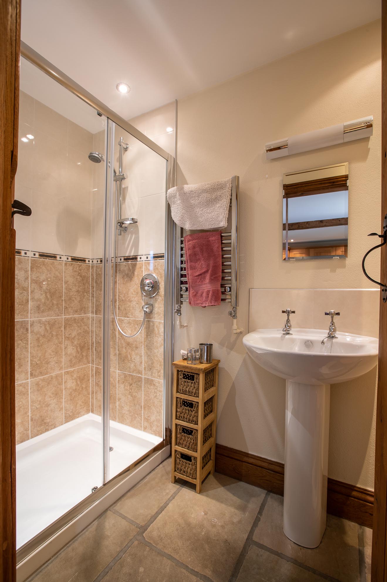Bathroom with shower, pedestal sink, towel warmer, and small wicker shelf. Beige and brown tones.