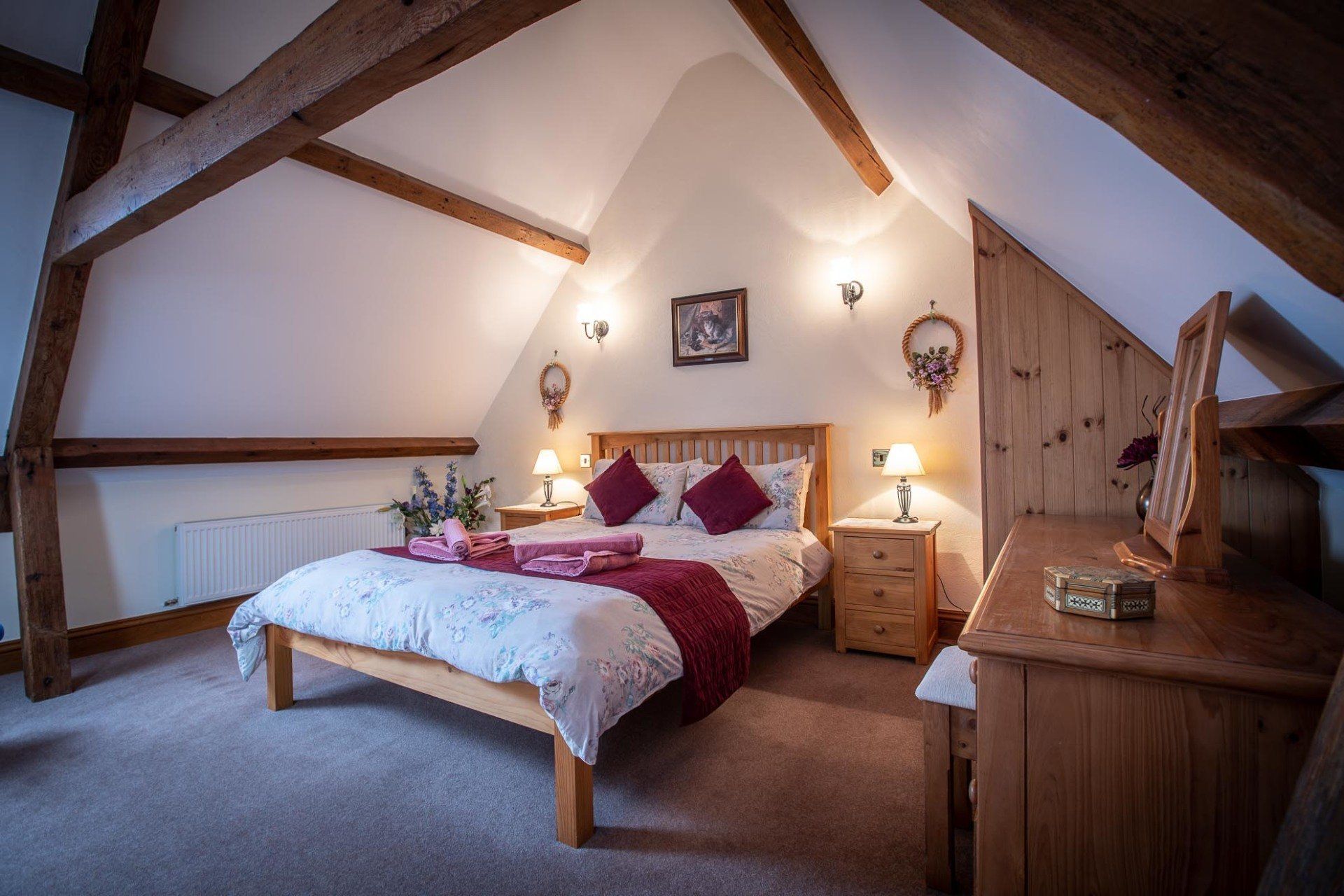 Bedroom with a wooden bed frame, beams, and dresser. White bedding, burgundy accents, and warm lighting.