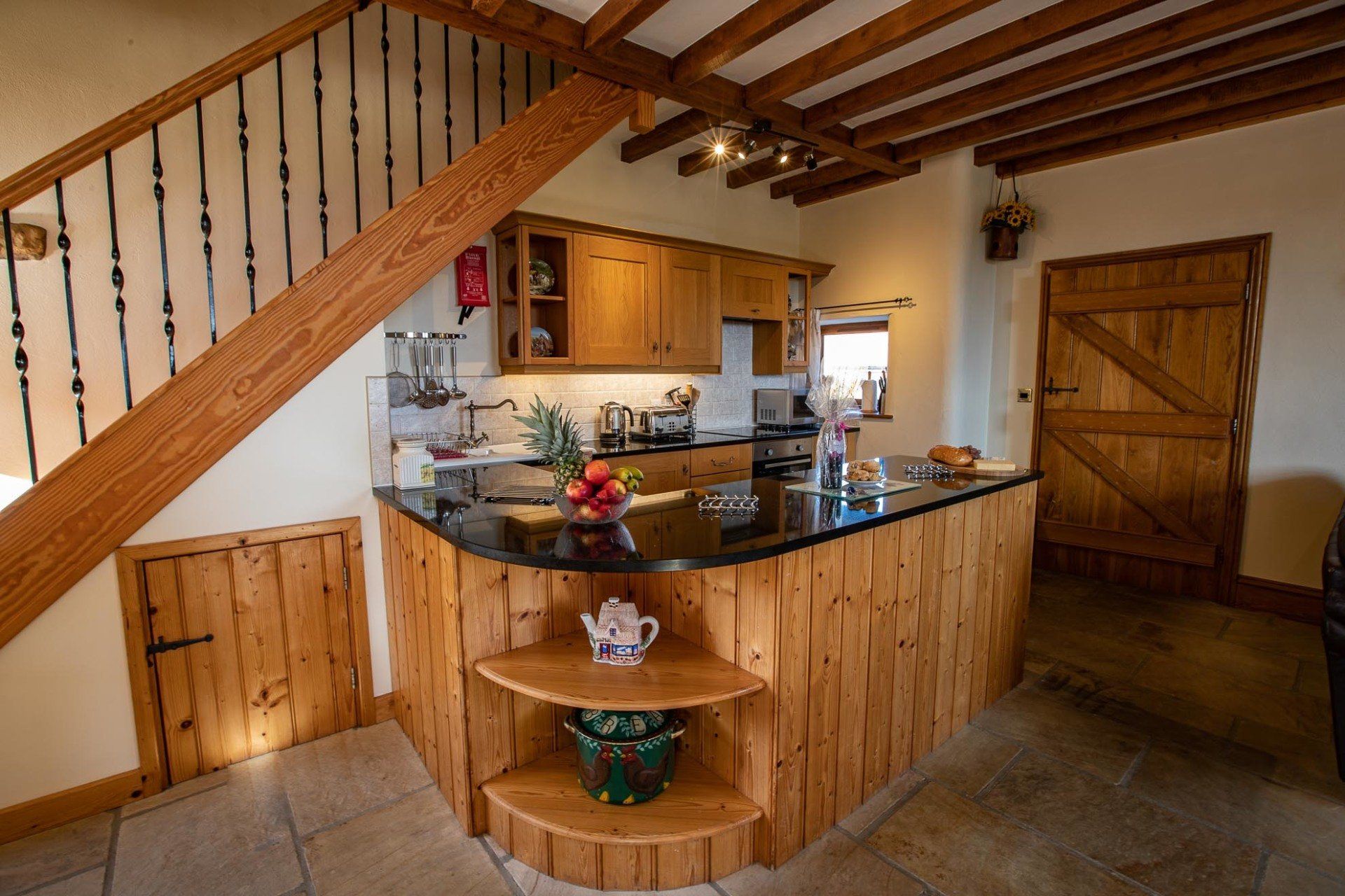 Kitchen with wooden cabinets, black countertop, and stone floor. A staircase and wooden door are visible.