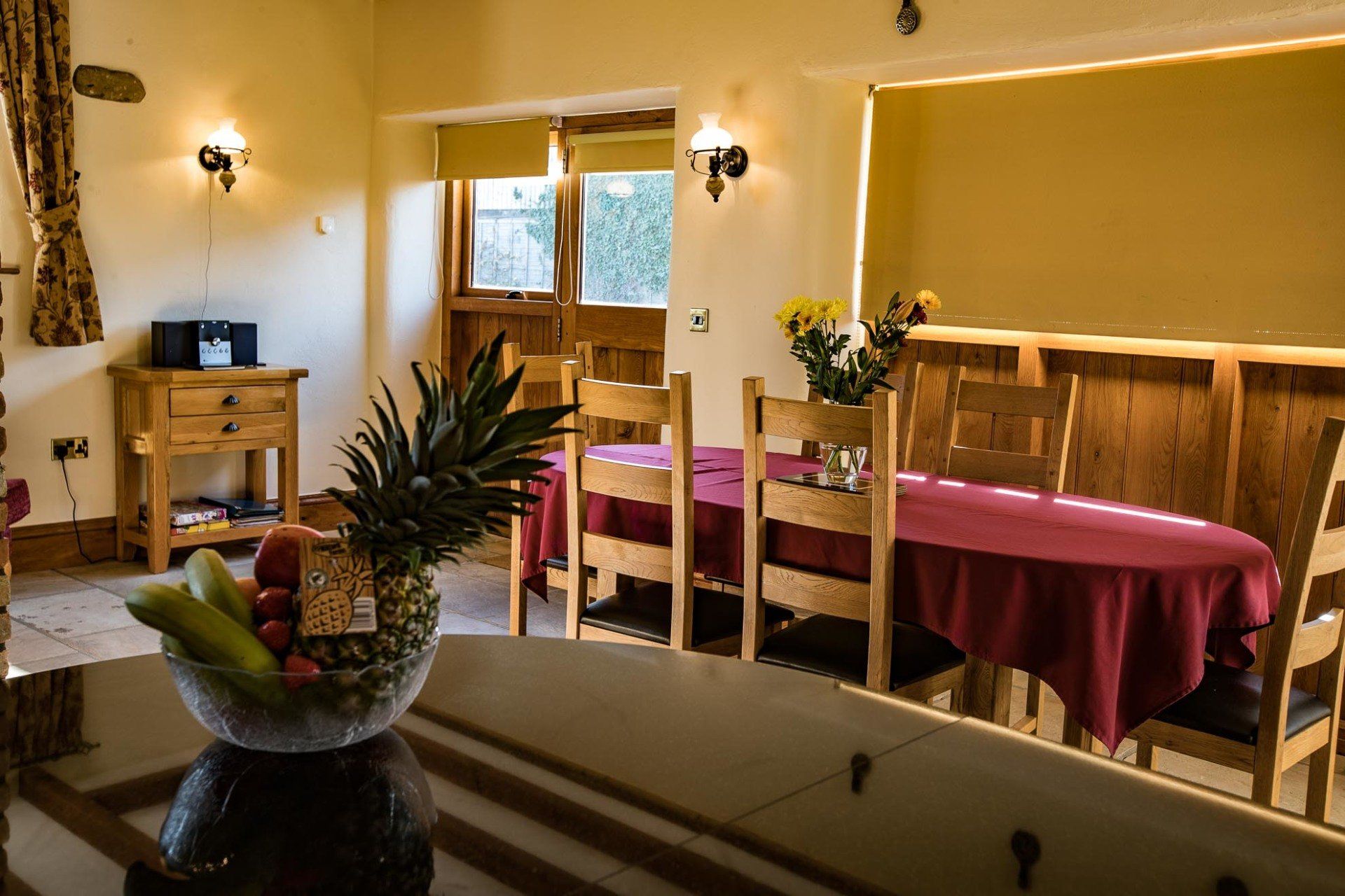 Dining room with wooden table, chairs, fruit bowl, and flowers. Sunlight streams through the window.