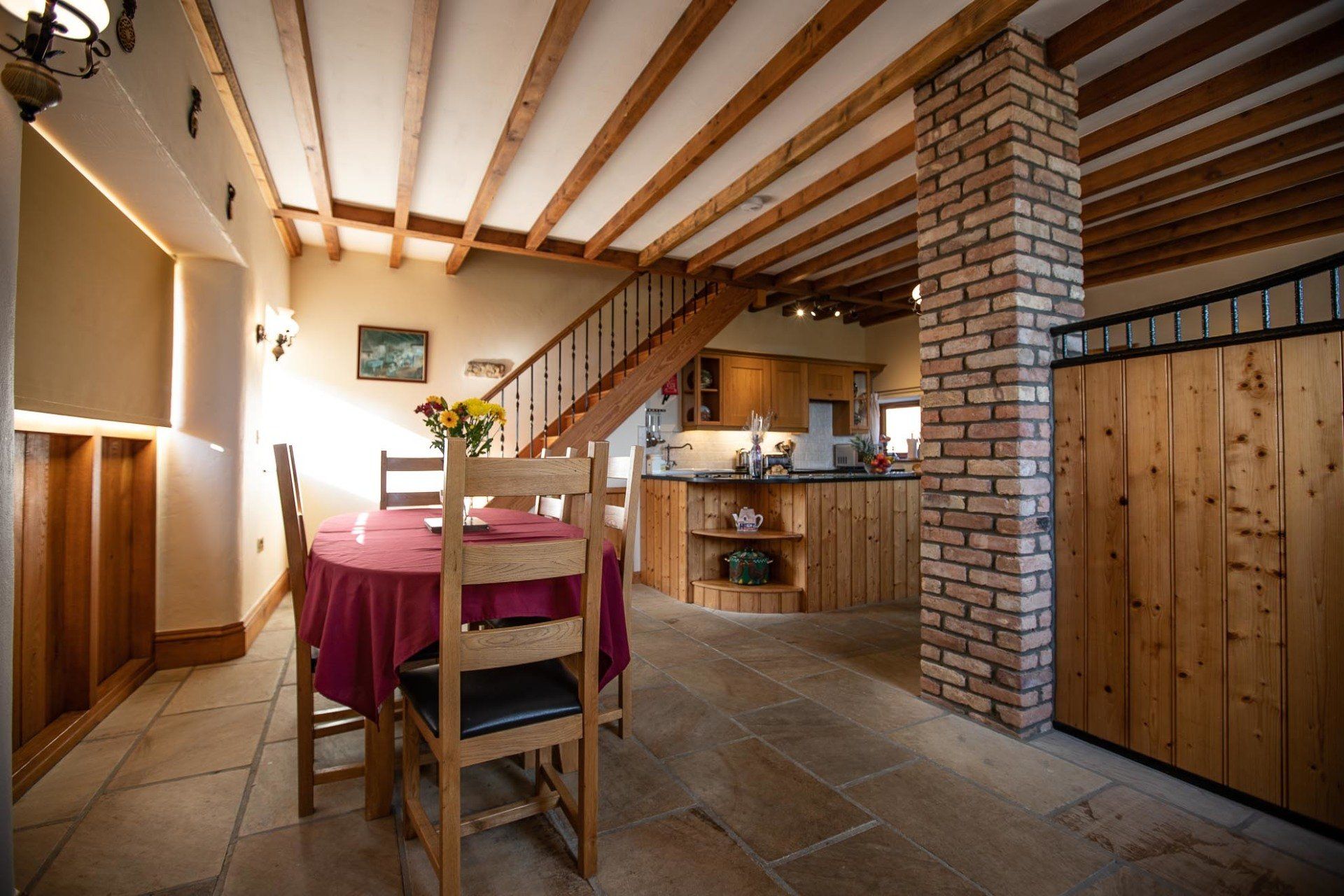 Rustic kitchen and dining area with wooden beams, stairs, and brick pillar; a round table is set for dining.