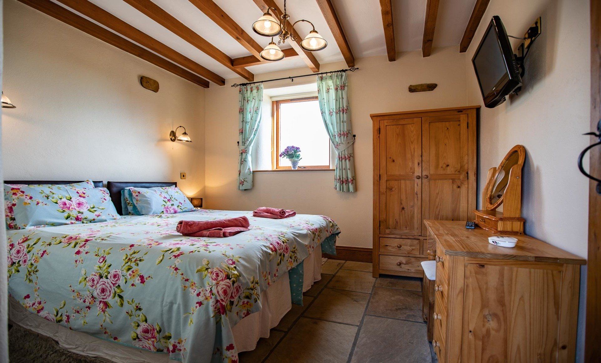 Bedroom with king-sized bed, floral bedding, wooden furniture, stone floor, and a small TV mounted on the wall.