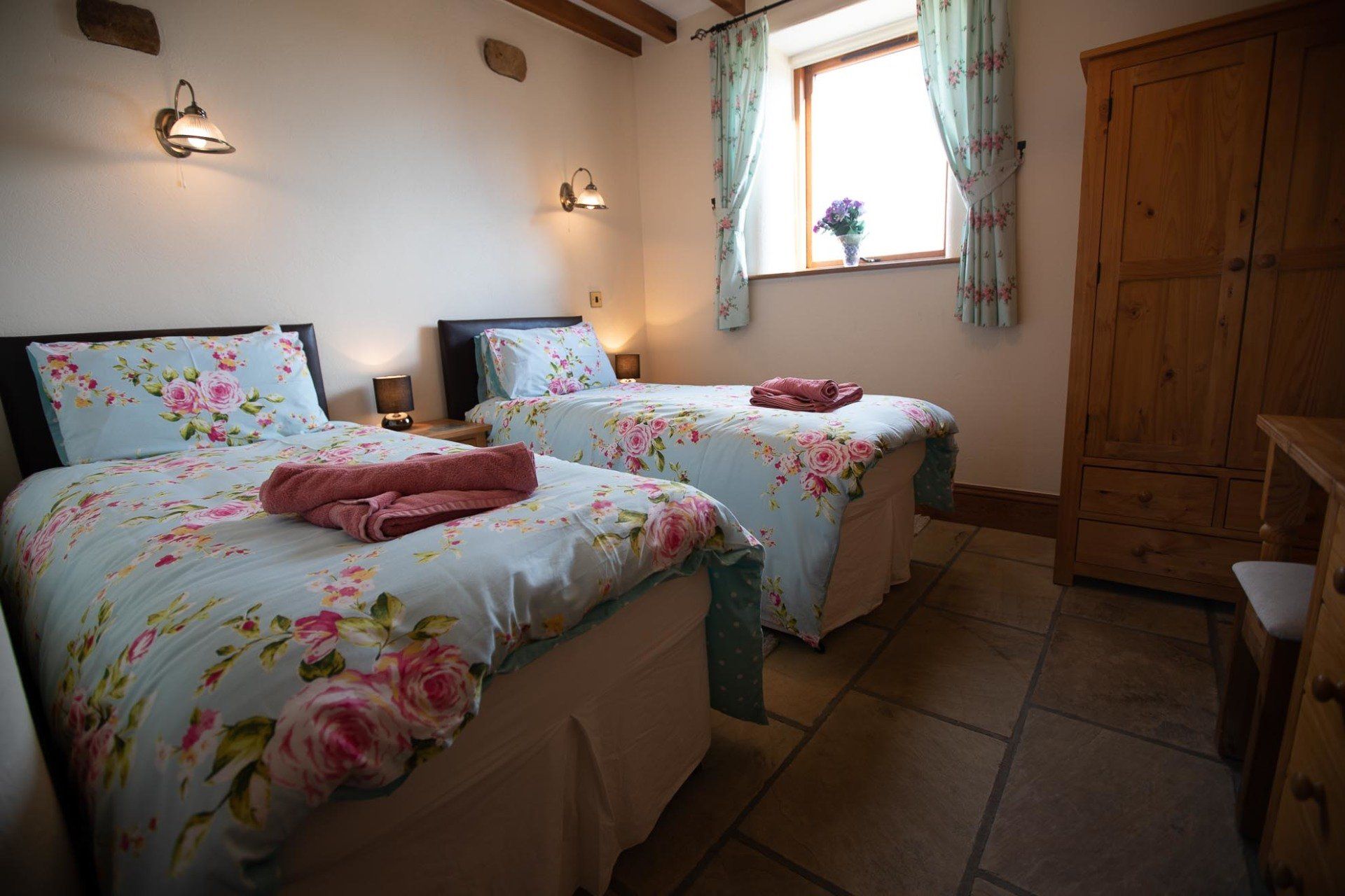Twin beds in a rustic bedroom, with floral bedding, a wooden wardrobe, and a window with curtains.