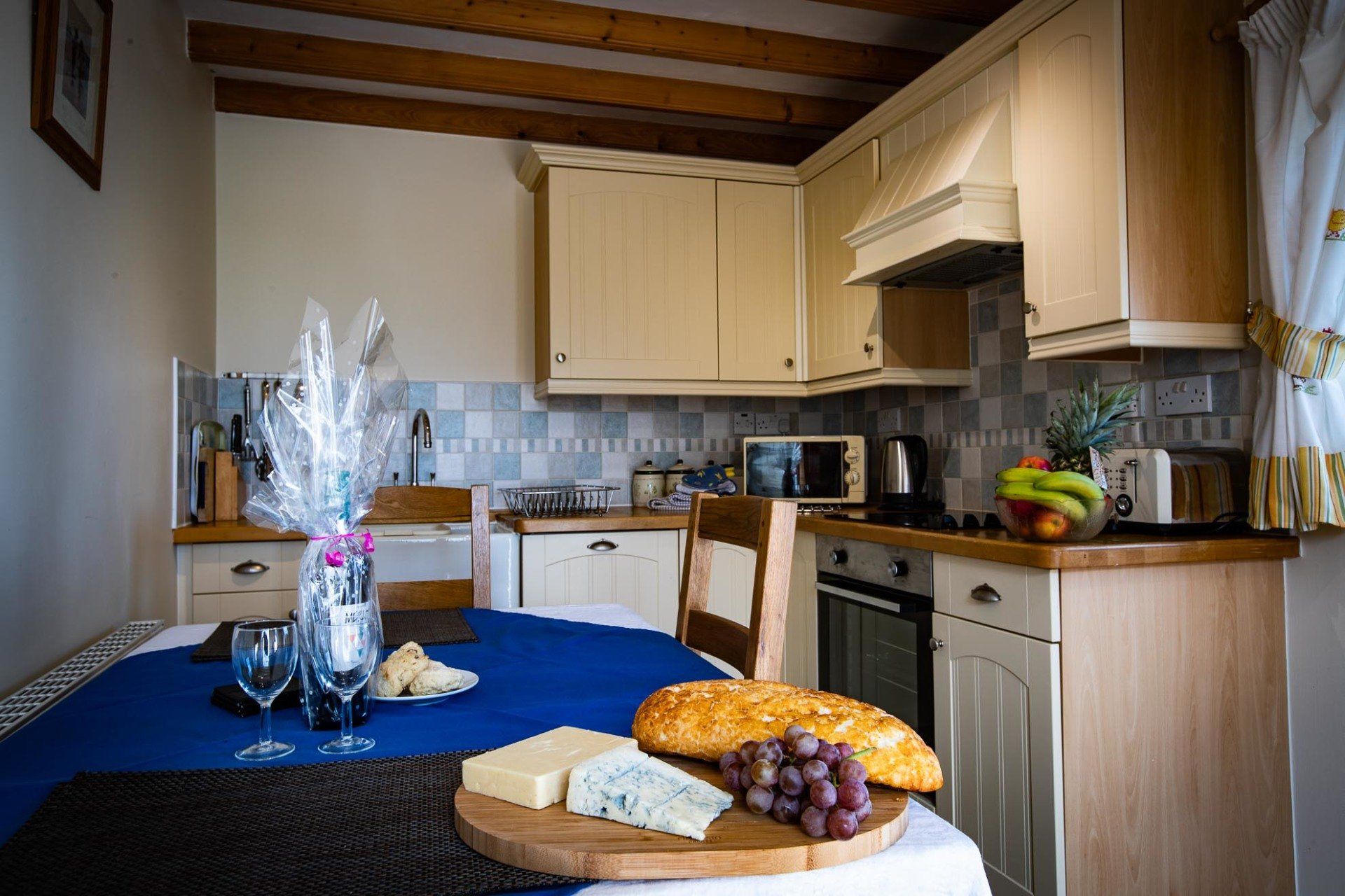 Cozy kitchen with blue tablecloth, food, and wine glasses. Light-colored cabinets, tiled backsplash, and wooden beams.