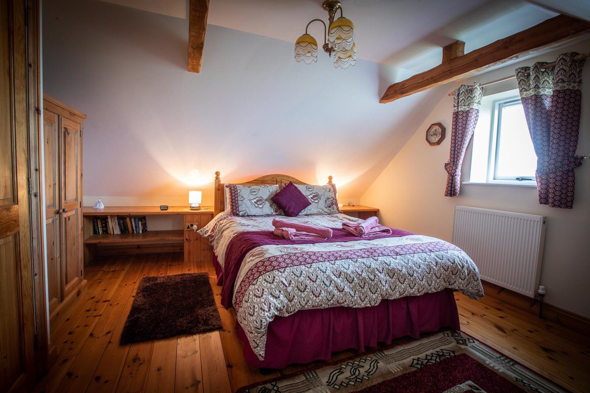 Bedroom with a bed, wooden floor, and a window with curtains. Warm lighting and a wardrobe.
