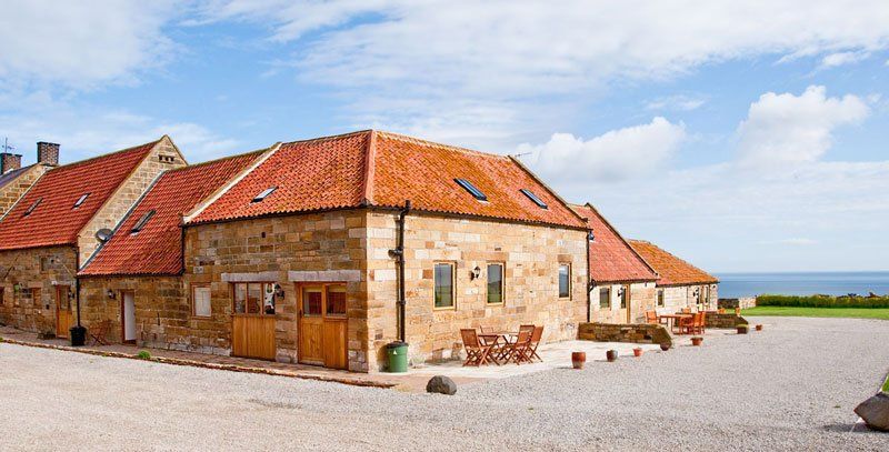 Stone buildings with orange roofs and a gravel courtyard, overlooking a blue ocean under a partly cloudy sky.