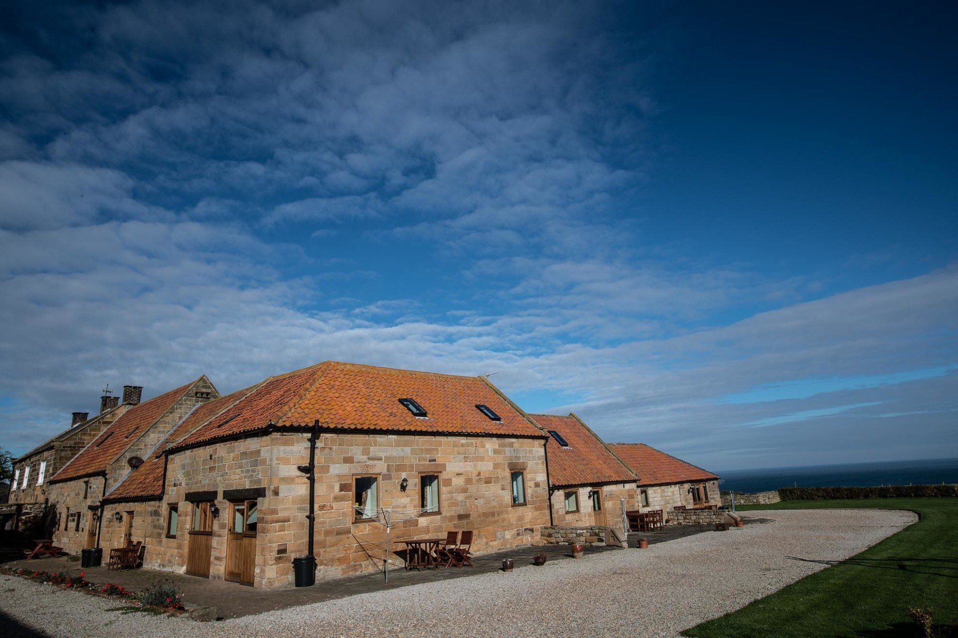 Stone cottages with orange tile roofs under a blue sky, gravel parking area, sea in background.