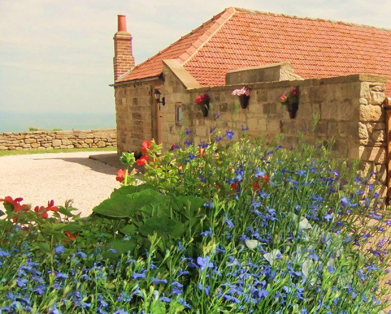 Stone building with red-tiled roof, flowers, and ocean view on a sunny day.
