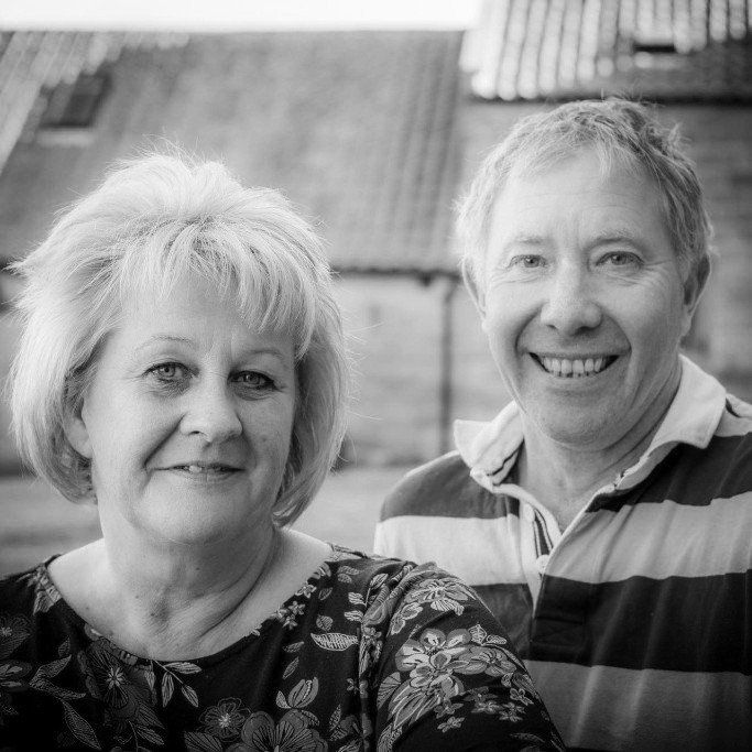 Smiling couple posing outdoors, woman with curly hair, man in striped shirt, old building in background.