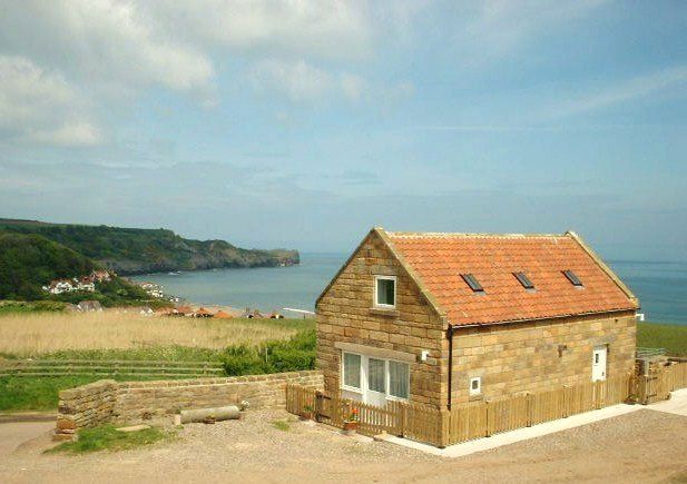 Stone cottage with orange tile roof, overlooking a beach and ocean on a sunny day.