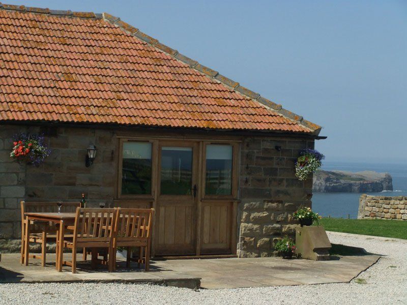 Stone cottage with brown tiled roof, wooden doors, and patio furniture overlooking the sea.