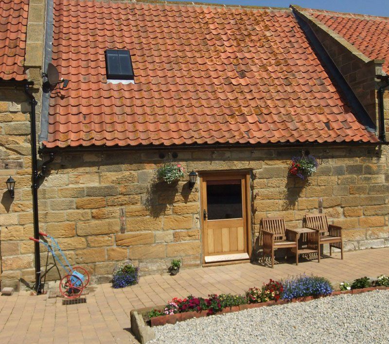 Stone cottage with red tiled roof, wooden door, small patio, and potted flowers.