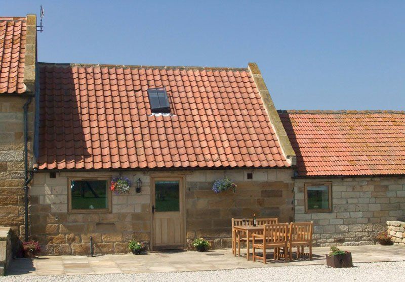 Stone cottage with red-tiled roof, wooden door, windows, and patio with table and chairs.