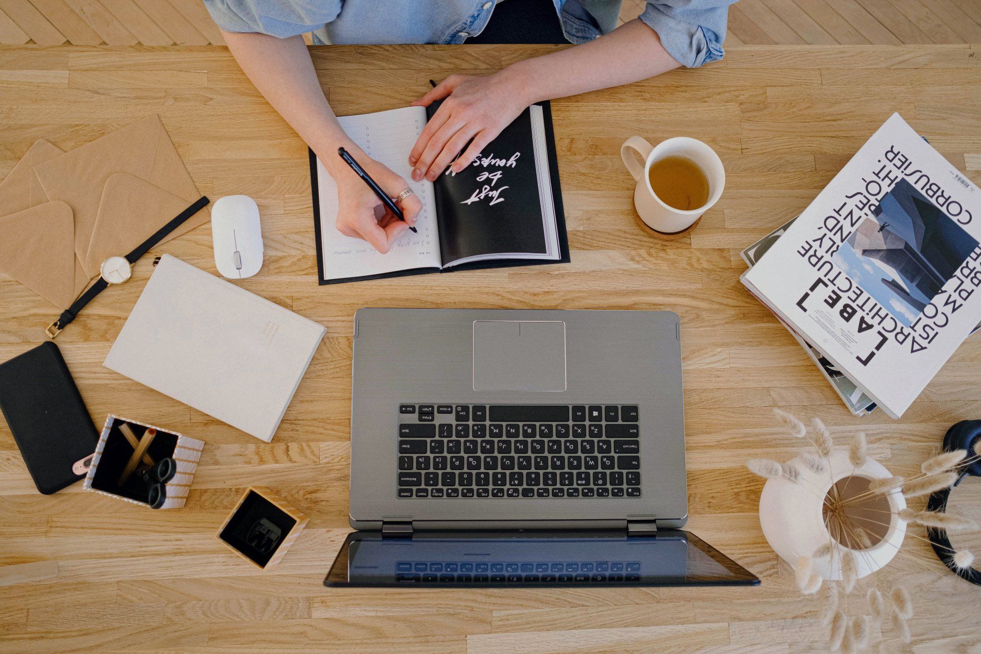 someone writing in a diary at their laptop with a mug of tea