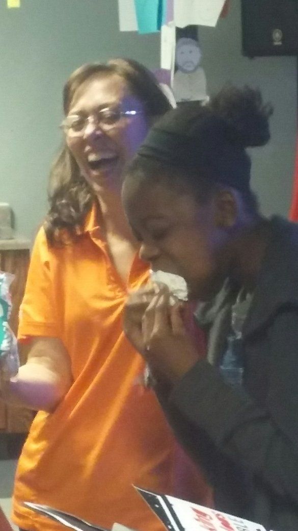 A woman in an orange shirt is standing next to a girl who is eating a piece of food