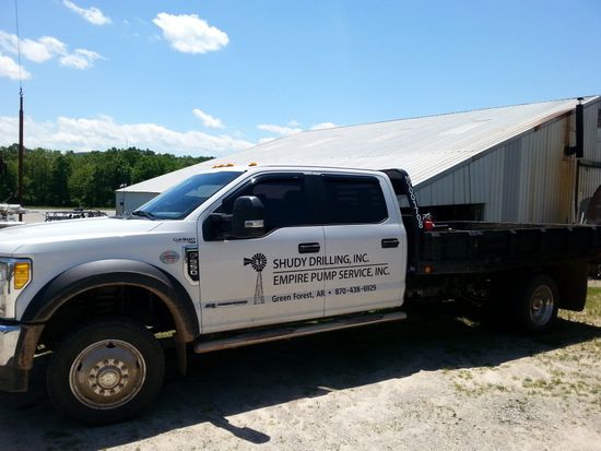 A white flatbed truck parked outside a metal industrial building under a sunny blue sky.