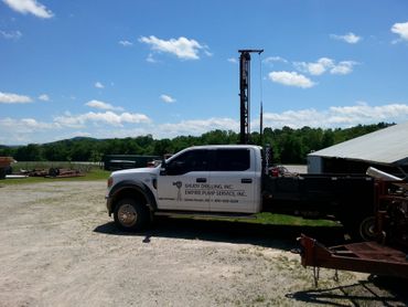 A white pickup truck with a mounted drilling rig parked on a gravel lot under a sunny blue sky.