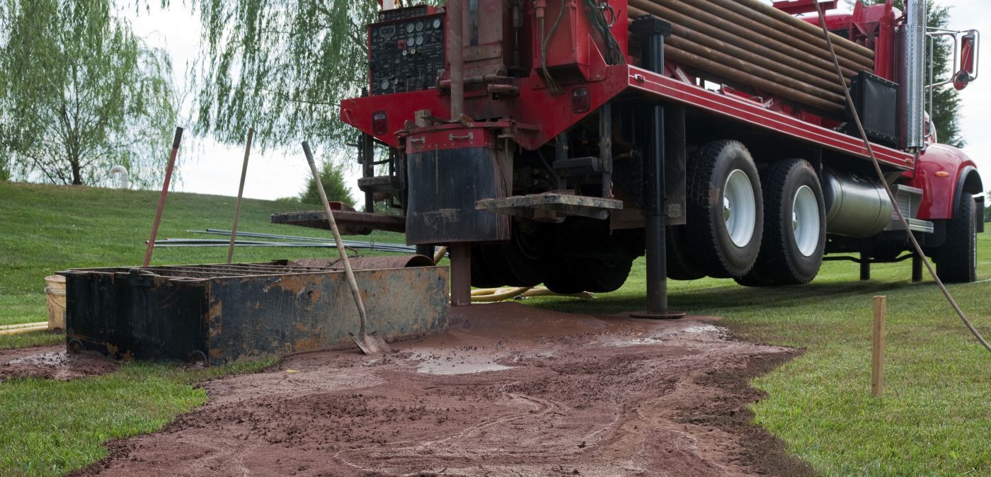 A red drilling truck parked on a grassy field, with mud and drilling equipment positioned over a hole in the ground.