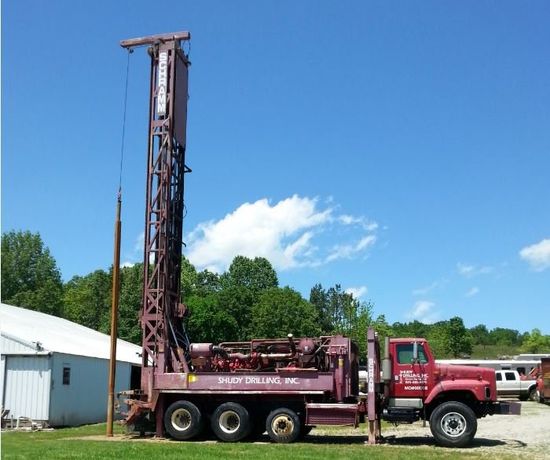 A red Schramm drilling rig truck with its mast raised, parked on a grass lawn near a building under a clear blue sky.