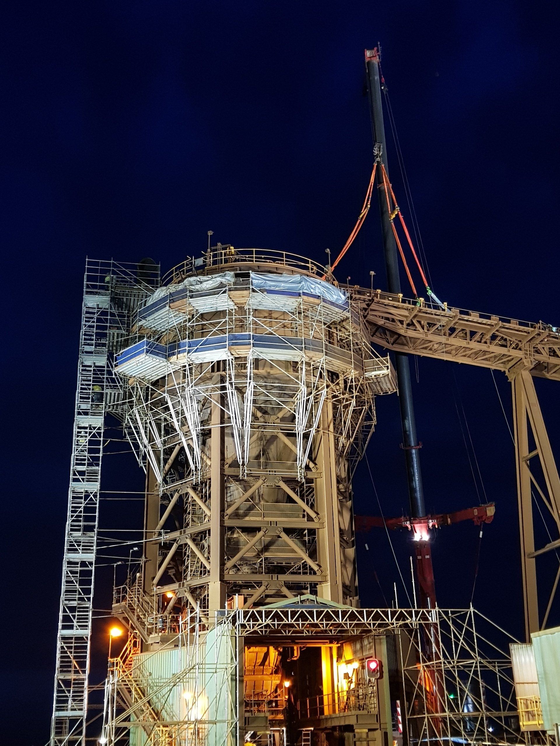 Lit industrial tower under construction at night, with cranes and bright work lights against a dark sky