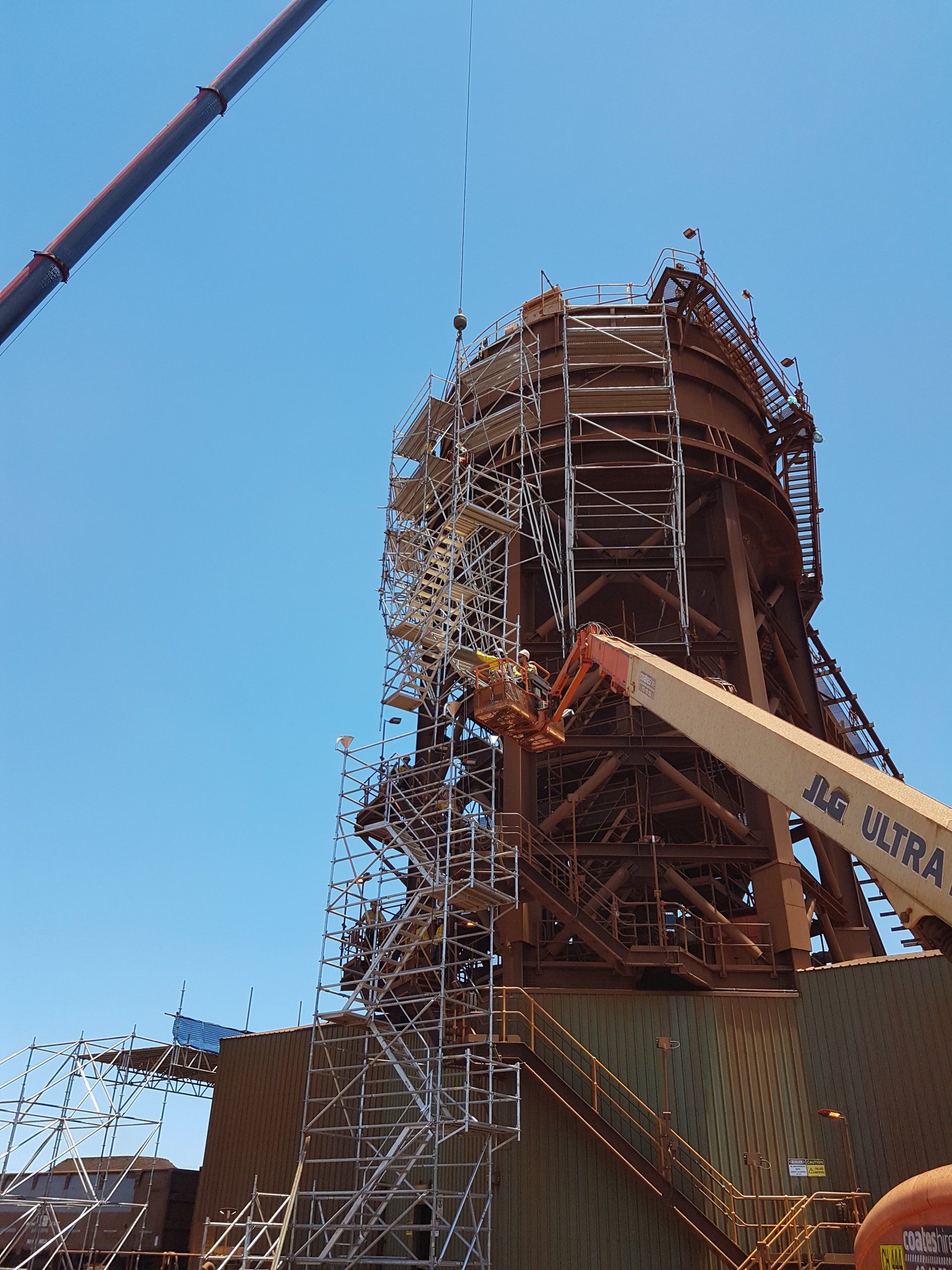 Large rust-colored industrial structure with scaffolding under a clear blue sky, with a crane boom nearby