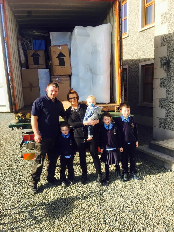 Family of six poses with a moving truck, boxes, and furniture. Bright day, stone house in the background.