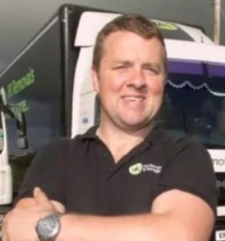 Man with crossed arms, smiling in front of a delivery truck. He wears a black shirt, watch. Truck has green branding.