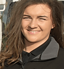 Young woman with brown hair smiling, wearing a black collared shirt and jacket, outside.