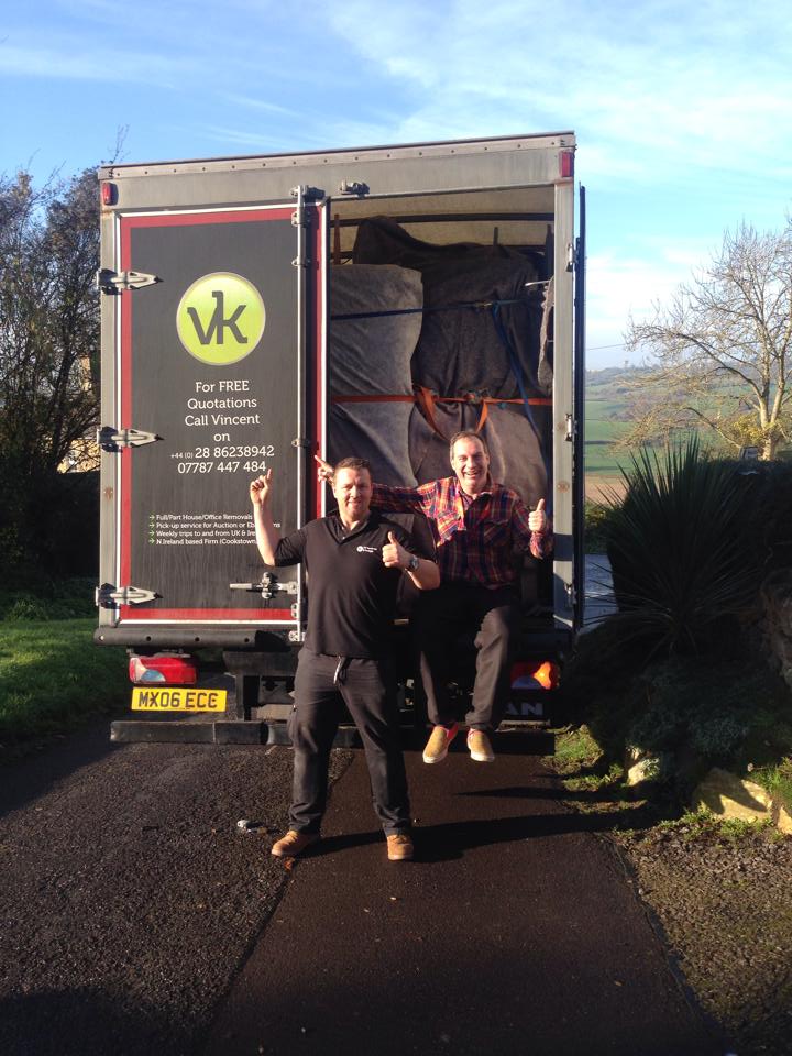 Two men smile, standing in the back of a truck. The truck is full of boxes. They are on a driveway in front of a green field.