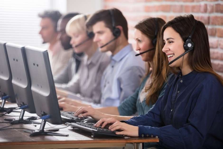 Um grupo de pessoas usando fones de ouvido estão sentadas em frente a monitores de computador em um call center.