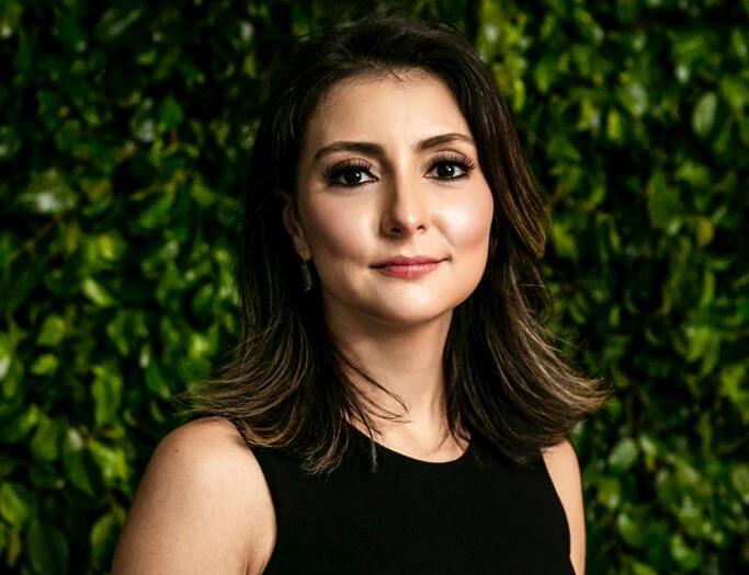 Uma mulher de vestido preto está parada em frente a uma cerca viva verde.