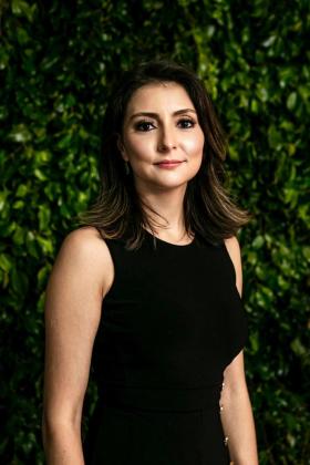Uma mulher de vestido preto está parada em frente a um arbusto verde.