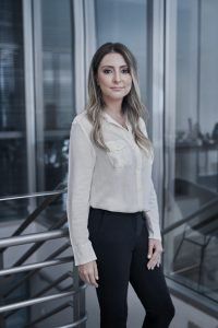 Uma mulher de camisa branca e calça preta está parada ao lado de uma parede de vidro.