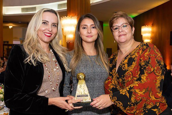 Três mulheres estão de pé, uma ao lado da outra, segurando um troféu.