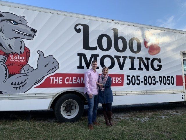 Couple standing in front of a Lobo's Moving truck with a wolf logo, thumbs-up pose, and phone number.