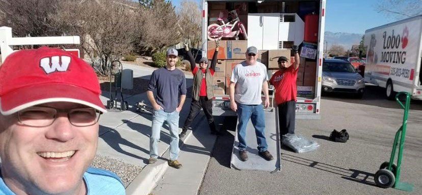 Group of movers unloading a truck, smiling man in a red hat takes a selfie, outdoor scene.