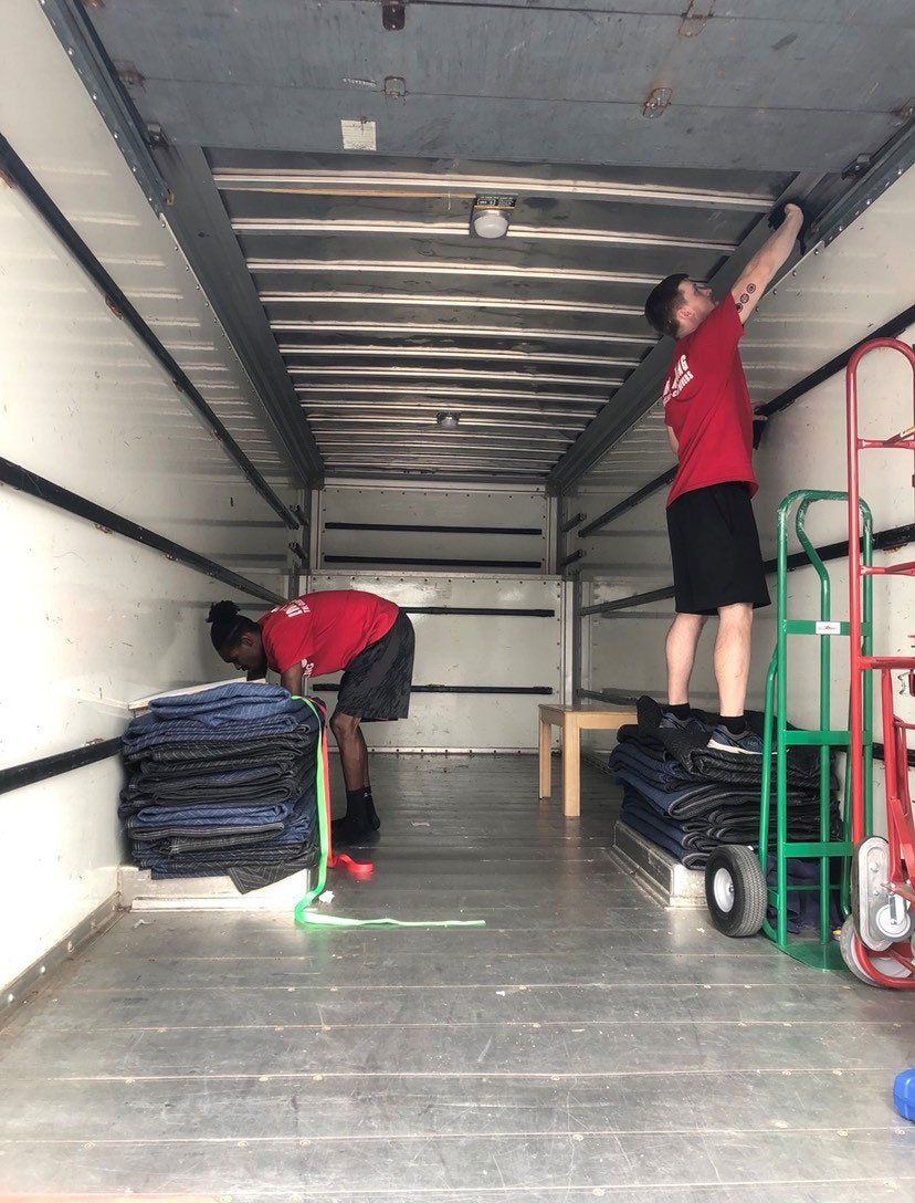 Two men in a moving truck load boxes, one on a dolly and one reaching overhead.