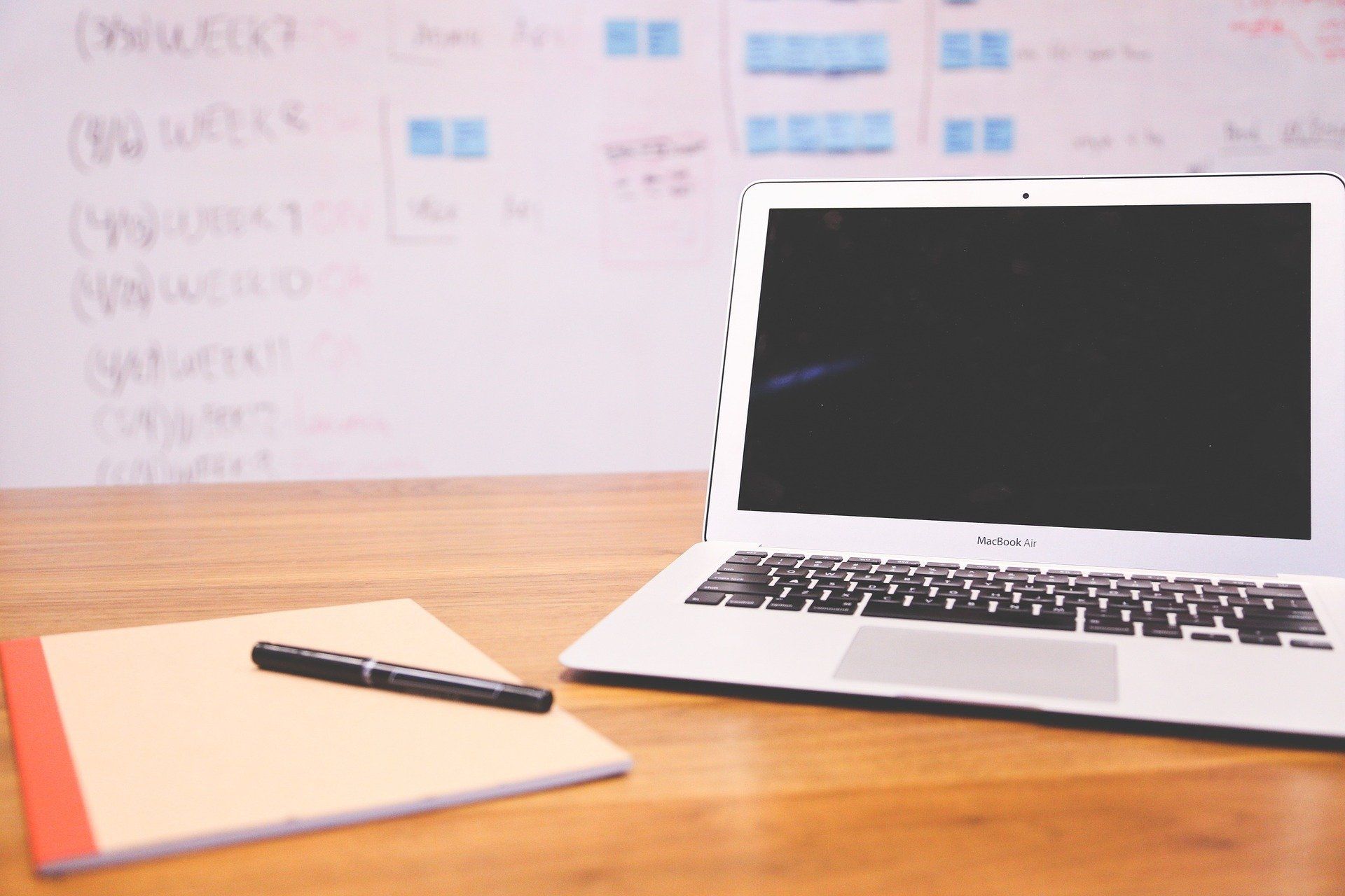 A laptop is sitting on a wooden table next to a notebook and pen.