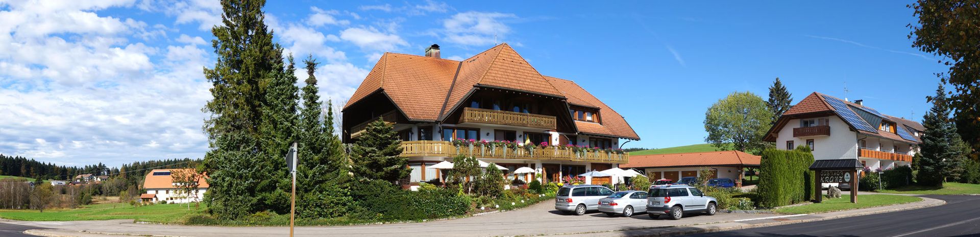 Ein großes Haus mit einem roten Ziegeldach ist von davor geparkten Autos umgeben.